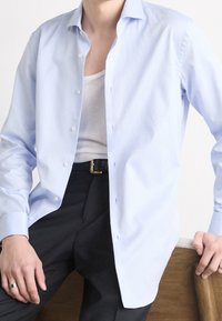 Světle modrá košile s dlouhým rukávem a knoflíky, s texturovaným vzorem, klasickým límcem a bílými knoflíky, doplněná tmavými kalhotami a opaskem.