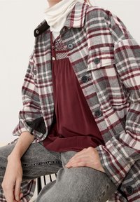 Grau, weiß und burgunderrot kariertes Jackett mit schwarzen Knöpfen. Darüber ein kastanienbraunes Oberteil und graue Jeans. Glatte Textur mit lockerer Passform.