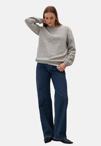 Szary sweter z dzianiny z długimi rękawami, okrągłym dekoltem i ściągaczem, zestawiony z ciemnoniebieskimi spodniami o szerokich nogawkach. Modelka stoi z jedną ręką na głowie.