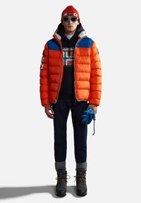Chaqueta acolchada naranja brillante con detalles en azul, superpuesta sobre un sudadera negra con capucha. Guantes azules, vaqueros oscuros y botas negras completan el atuendo.