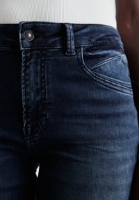 Temno modri kavbojki iz denima z gladko teksturo, ki imajo zaponko, sprednje žepi in subtilne izblitene podrobnosti na tkanini.