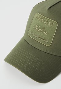 Olívazöld sapka, melyen egy varrott téglalap alakú folt található. A fóliázott szöveg olvasható: "Buffalo, NY" és "New Era Established 1920." Sima szövet textúra.