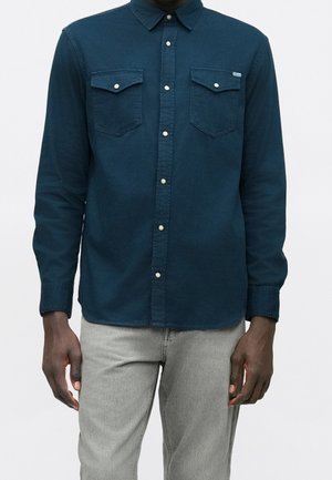 Homme portant une chemise bleu foncé à boutons avec deux poches poitrine et un pantalon gris clair sur un fond uni clair.