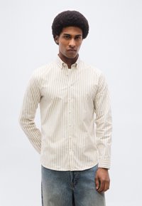 Jeune homme avec une coiffure afro portant une chemise à rayures verticales beige et blanche boutonnée et un jean bleu délavé, debout devant un fond uni.
