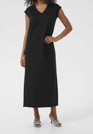 Femme portant une robe midi noire sans manches à col en V et des sandales à talons ouvertes à bout effiloché bleu clair, debout, une main sur la hanche.
