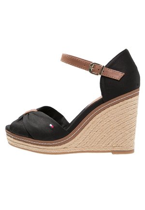 Schwarze Keilsandalette mit geflochtenem Plateauabsatz, braunem Knöchelriemen mit Schnalle und vorderer Knotendetail aus Stoff mit kleinem Flaggenmotiv.