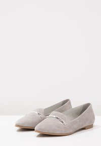 Grijze suède ballerina's met een spitse neus, voorzien van een glanzend metallic accent over de wreef, geplaatst op een witte ondergrond.