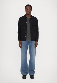 Schwarze button-up Jacke mit zwei Brusttaschen, graues Crew-Neck-T-Shirt, helle, weit geschnittene Jeans und schwarze Schuhe. Einfaches, strukturiertes Design.