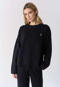 Maglione nero lavorato a maglia con orlo e polsini a coste. Presenta una scollatura rotonda e un dettagliato logo discreto sul petto. Vestibilità casual e rilassata.