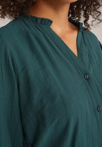 Teal Bluse mit Knopfleiste aus strukturiertem Stoff, mit Rüschenkragen und vertikalen Streifen, entspannter Passform und drei sichtbaren Knöpfen.