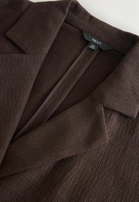 Brun blazer i et tekstureret stof, med et spidset revers og åben front. Etiket synlig ved halsudskæringen, med lige skåret ærmer.