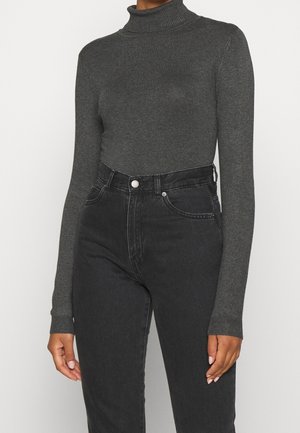 Personne portant un pull col roulé ajusté gris foncé et un jean noir taille haute sur un fond uni.