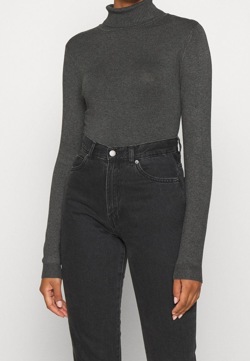 Personne portant un pull col roulé ajusté gris foncé et un jean noir taille haute sur un fond uni.