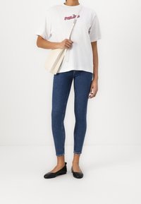 Bílý bavlněný tričko s červeným a modrým textem, v kombinaci s tmavě modrými skinny džíny a černými balerínami. Krémová crossbody taška s úzkým designem.