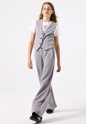 Chaleco gris ajustado con cierre de botón, llevado sobre una camiseta blanca, combinado con pantalones sueltos de cuadros grises y zapatos negros. Atuendo simple y coordinado.