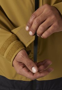 Manche de veste jaune avec un poignet ajustable en Velcro. Des mains ajustent le poignet, révélant une texture lisse et une section inférieure foncée contrastante.