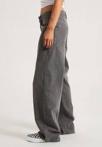 Graue weit geschnittene Jeans mit sichtbaren Stickdetails, mittelhoher Taille und großen seitlichen Taschen. Getragen mit schwarz-weißen Kariertenschuhen zum Hineinschlüpfen.
