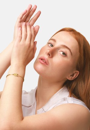 Bracciale d'oro con ciondolo rotondo, dal design a catena, indossato su un polso. La modella ha unghie rosa chiaro, pelle liscia e lunghi capelli rossi.