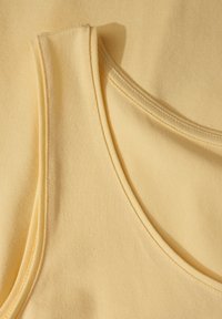 La camiseta sin mangas de algodón amarillo claro presenta un escote redondeado y tirantes anchos. Textura suave con detalle de costura visible a lo largo de los bordes.