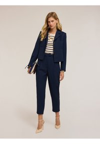 Completo da uomo blu navy composto da un blazer corto con due bottoni, pantaloni su misura e una maglietta a righe, abbinato a tacchi alti di colore chiaro.