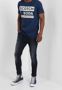 Navyblaue Baumwoll-T-Shirt mit weißem Grafiktext, kombiniert mit dunkelblauen Slim-Fit-Jeans und blauen High-Top-Sneakern mit weißen Akzenten.