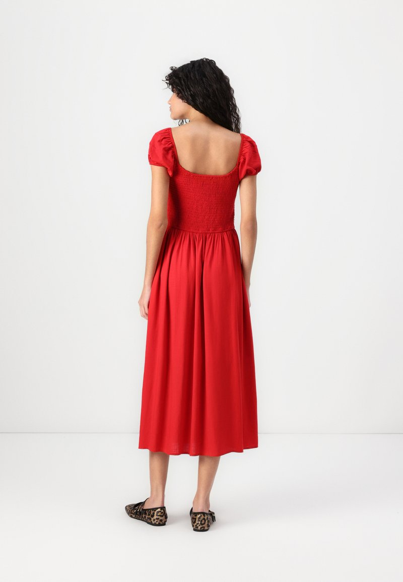 Rotes Kleid mit kurzen Puffärmeln, gesmoktem Oberteil und fließendem Rock. Der Stoff hat eine glatte Textur. Getragen mit flachen Schuhen im Leopardenmuster.