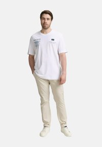 Witte t-shirt met tekstontwerpen in blauw en zwart, gecombineerd met licht beige broek en witte sneakers. Eenvoudige, ontspannen pasvorm, korte mouwen.