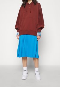 Sudadera con capucha color burdeos y bolsillo frontal, superpuesta sobre una falda azul con pliegues. Zapatillas blancas con detalles negros y calcetines cortos.