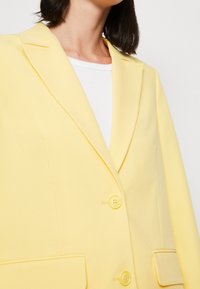 Żółta marynarka wykonana z gładkiego materiału, z kołnierzem z wycięciem, dwoma guzikami oraz przednimi kieszeniami z lamówką.