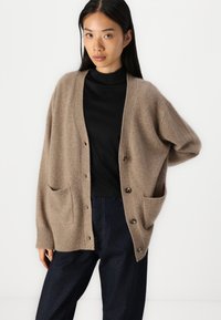 Beige pletený cardigan s volným střihem, s výstřihem do V, velkými knoflíky a dvěma předními kapsami. Nosený přes černý top.