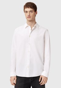 Camisa blanca de manga larga con cuello clásico y pequeño detalle bordado en el pecho. Tela suave, corte estándar y textura ligera.