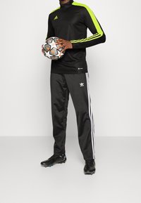 Svart långärmad atletisk tröja med neongröna ränder, kombinerad med svarta sweatpants med vita sidostripes och Adidas-logotyp, håller en fotboll.