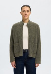 Olivno zelena jopa iz mehke tkanine, z visokim ovratnikom, odprtim sprednjim delom in dvema sprednjima žepoma. Nošena preko svetlega topa in kavbojk.