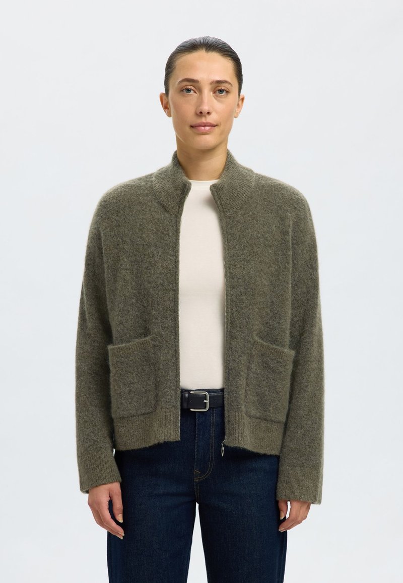 Olivno zelena jopa iz mehke tkanine, z visokim ovratnikom, odprtim sprednjim delom in dvema sprednjima žepoma. Nošena preko svetlega topa in kavbojk.