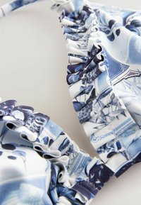 Top bikini con bordi arricciati, caratterizzato da un motivo blu e bianco con disegni floreali e scenici, realizzato in tessuto elasticizzato.