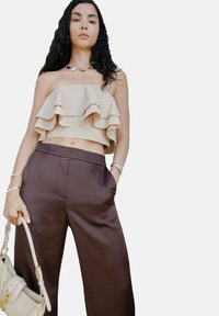 Haut court beige à volants avec des fronces superposées, associé à un pantalon large marron. Les accessoires incluent un collier en argent et un sac à main.