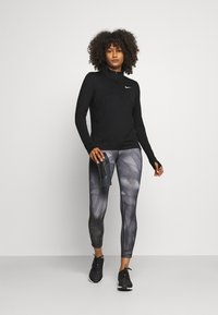 Black half-zip long-sleeve top paired with gray abstract-patterned leggings. Model holds a black water bottle. Black athletic shoes.