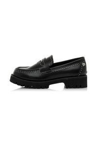 Mocassin en cuir noir avec semelle épaisse, détails en clous argentés le long des bords et un petit logo métallique sur le talon.