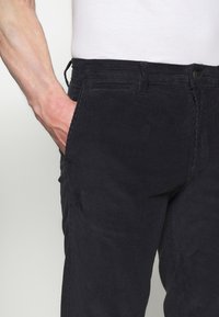 Černé kordové kalhoty s texturovanou tkaninou, slim fit, boční kapsy a knoflíkovým zapínáním. Je viditelný nastavovací stahovací pas.