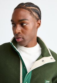 Green fleece jacket with cream collar, featuring a zip closure and button accents. Worn over a white shirt, textured fabric visible.