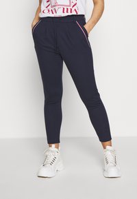 Calças jogger azul-marinho com cintura elástica, bolsos frontais e riscas laterais contrastantes em branco e vermelho, combinadas com ténis brancos de solado grosso.