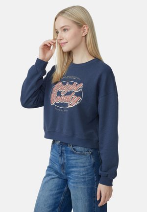 Ung kvinde med langt blond hår iført en navy sweatshirt med teksten "Bright Beauty" og blå jeans, smilende, stående mod en hvid baggrund.