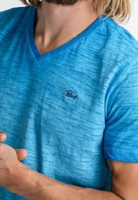 Blå V-ringad t-shirt med en marmorliknande struktur. Har en liten svart logotyp på bröstet, korta ärmar och en avslappnad passform.