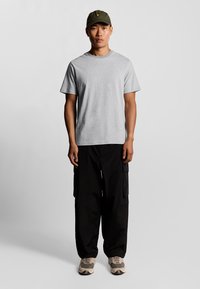 T-shirt in cotone grigio, abbinato a pantaloni cargo neri. Cappellino verde oliva con logo giallo. Le scarpe presentano un design beige e nero.