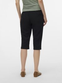 Cilvēks valkā melnas īsās bikses, zaļu ribotu topu un brūnas plakanas kurpes, stāvot uz balta fona.