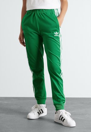 Osoba nosząca zielone spodnie dresowe Adidas z białymi paskami i białe buty sportowe Adidas stojąca na szarej podłodze.