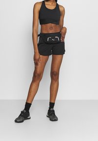 Idrottslig person klädd i svart sport-bh, shorts, svarta sneakers och en svart Puma midjeväska, stående framför en enfärgad vit bakgrund.