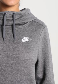 Grå sweatshirt med hög krage, snöre för justering och en vit Nike-logotyp på bröstet. Tillverkad av mjukt tyg med en slät textur.