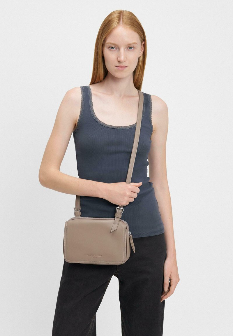 Borsa a tracolla in pelle beige con finitura liscia, chiusura con zip e tracolla regolabile, sostenuta contro una maglietta nera e jeans neri.