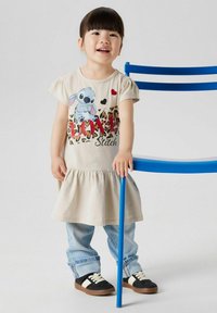 Jasno-beżowa sukienka z rozkloszowanym dołem, z grafiką Stitche'a i napisem "LOVE" w kolorze czerwonym z przodu. Połączona z niebieskimi dżinsami i czarnymi butami.